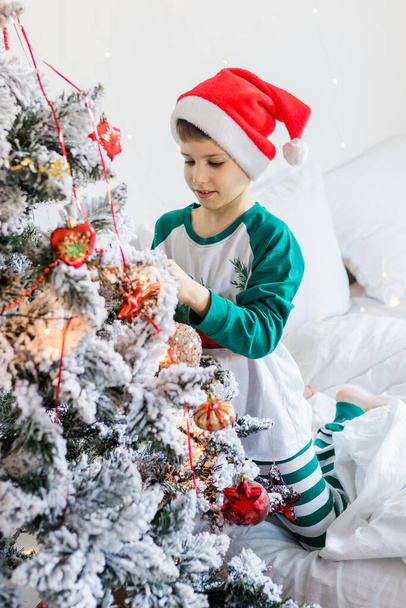 Το παιδί στολίζει το χριστουγεννιάτικο δέντρο. Το αγόρι κρατάει ένα χριστουγεννιάτικο παιχνίδι. Πρωί πριν τα Χριστούγεννα στο σπίτι. Γενιά άλφα και γονιδίων άλφα παιδιά - Φωτογραφία, εικόνα