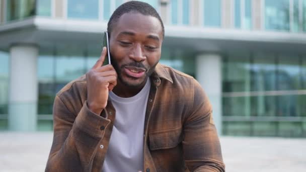 Afrikkalainen amerikkalainen mies puhuu älypuhelimella kadulla kaupungin ulkona. Mies, jolla on kännykkä, juttelee ystävien kanssa. Hymyilevä henkilö soittaa kännykällä. Mies, jolla on keskustelu matkapuhelimeen - Materiaali, video