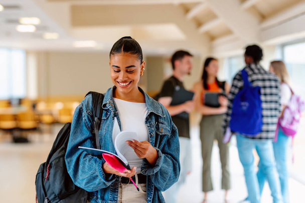 Retrato de una chica latina sonriente mirando sus notas de la universidad en un pasillo en el campus o la escuela secundaria con sus compañeros de clase en el fondo. Tiempo de examen. - Foto, imagen