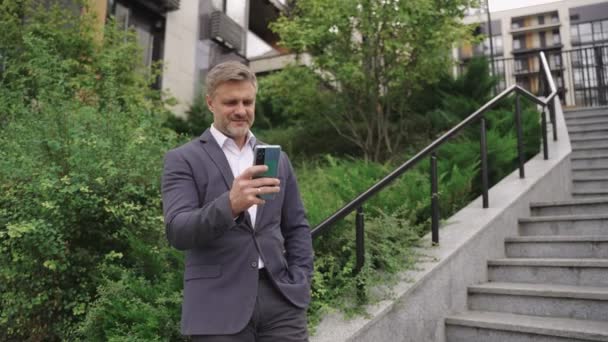Усміхнений бізнесмен, використовуючи смартфон, стоїть біля сходів у місті. Бізнес, комунікація, концепція технології. Повільний рух - Кадри, відео