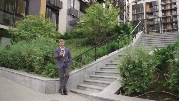 Boldog üzletember mobiltelefont használ, miközben a szabadban pózol a lépcsők közelében. Üzlet, kommunikáció, technológiai koncepció. Lassú mozgás. - Felvétel, videó