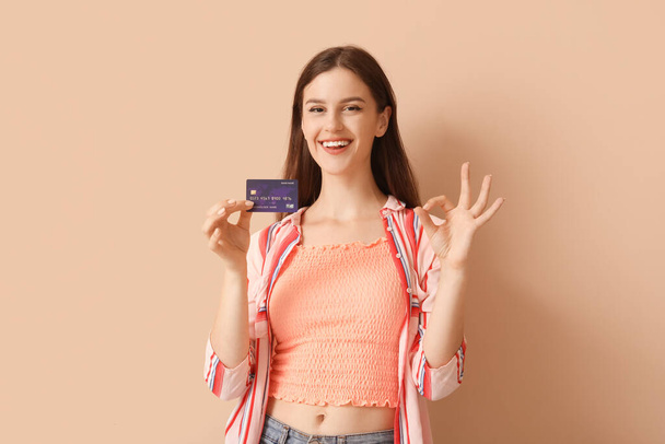 Νεαρή γυναίκα με πιστωτική κάρτα που δείχνει OK σε μπεζ φόντο - Φωτογραφία, εικόνα