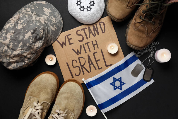 Tekst STAND With ISRAEL, mundur wojskowy, nieśmiertelniki, żydowski kapelusz, flaga Izraela i świece na ciemnym tle - Zdjęcie, obraz