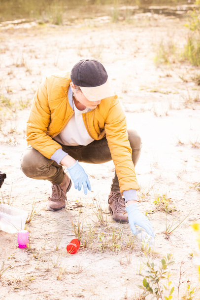 Esta foto captura la esencia de la responsabilidad ambiental. Una joven caucásica milenaria, equipada con una gorra, un traje de senderismo y guantes de goma, se ve arrodillada en la arena. Su enfoque - Foto, imagen