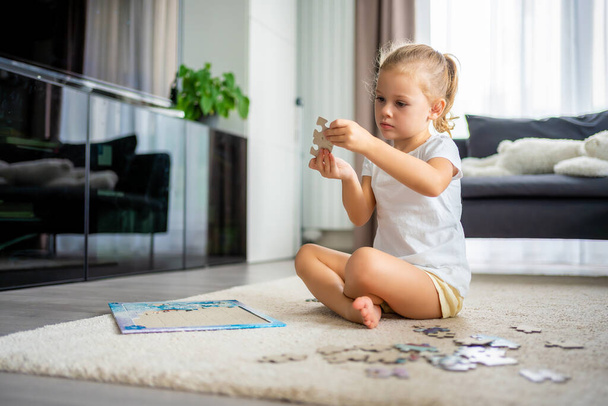 Little blonde girl sits at home on the carpet and collects puzzles. High quality photo - Photo, Image