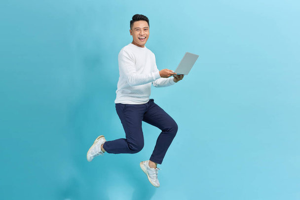 Веселый азиатский молодой человек держит ноутбук улыбаясь прыгая высоко работает изолированы на голубом фоне. - Фото, изображение