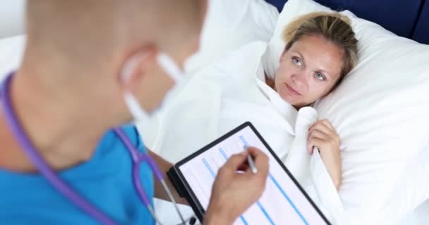 Lääkäri istuu sängyssä nuoren naisen potilaan ja kirjallisesti tietoa digitaalinen tabletti 4k elokuva. Säilyttäminen sähköisen sairaushistoria etänä käsite - Materiaali, video