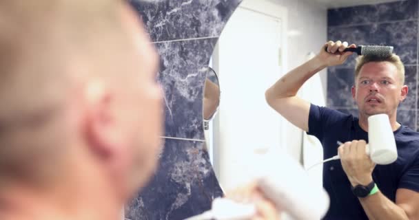 Молодой человек стоит у зеркала и расчесывает волосы феном в фильме в ванной комнате 4К. Уход за волосами для мужчин - Кадры, видео