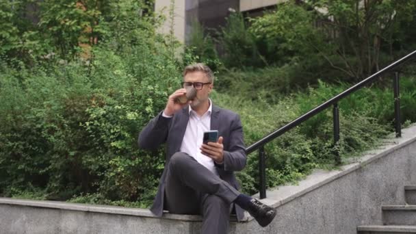 Un uomo d'affari sorridente che riposa bevendo caffè e usando lo smartphone all'esterno. Affari, comunicazione, concetto tecnologico. Rallentatore - Filmati, video
