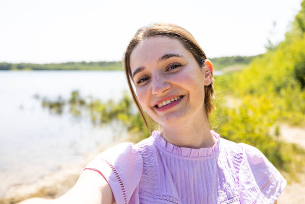 素晴らしいアウトドアの抱擁の中で,幸せな若い女性は湖畔でセルフィーをスナップし,日焼けした日の本質を捉える彼女の鮮やかな笑顔. このスナップショットは健康のお祝いです - 写真・画像