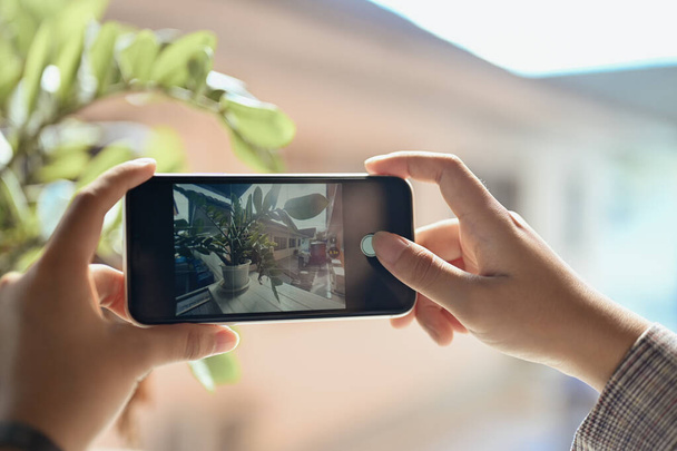 市内の晴れたバルコニーでポット植物の写真を撮る携帯電話を使用している女性. - 写真・画像