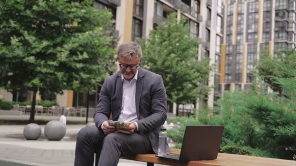 Dospělý obchodník s brýlemi sedí na lavičce a počítá peníze a pracuje s laptopem. Obchod, platební koncept. Zpomalený pohyb - Záběry, video