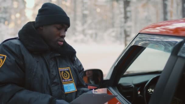 Mittlere Aufnahme eines afroamerikanischen Straßeninspektors in warmer Uniform, der mit Ordner neben orangefarbenem Auto steht und in Walkie-Talkie spricht - Filmmaterial, Video