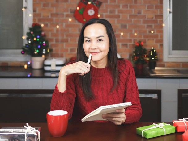 Portrét asijské ženy v červeném pleteném svetru sedí u stolu s červeným šálkem kávy a dárkové krabice v kuchyni s vánoční výzdobou, pomocí pera psaní seznamu jmen na notebooku.  - Fotografie, Obrázek