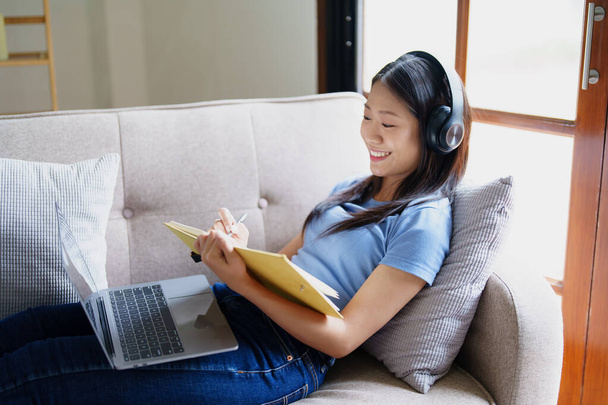 mulher usando fones de ouvido para ouvir música e ler notebook no sofá - Foto, Imagem