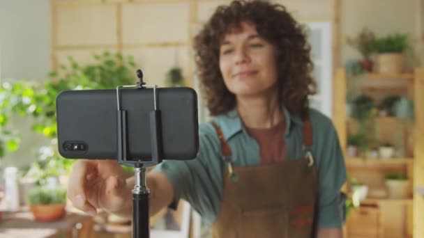 Enfoque en rack de seguidores de saludo jardinero mientras hace video sobre plantas de interior usando teléfono móvil en trípode en interiores - Imágenes, Vídeo