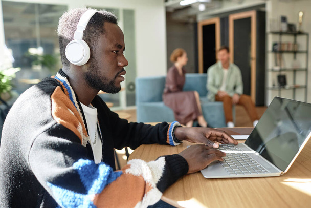 Afro-Amerikaanse zakenman in draadloze hoofdtelefoon werkt online op laptop tijdens het zitten in kantoor hal - Foto, afbeelding