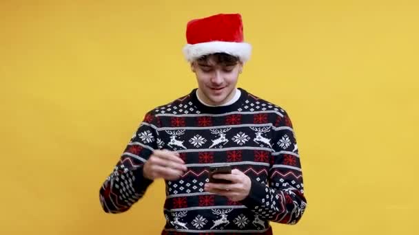 Kafkasyalı genç adam Noel Baba şapkası takıyor. Arka planda izole edilmiş başarılı bir ifadeyle cep telefonuyla yazı yazıyor. - Video, Çekim