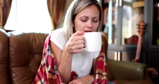 Mooie vrouw in geruite drankjes uit beker zittend op de bank. Warme thee of thee voor koorts en hoge lichaamstemperatuur - Video