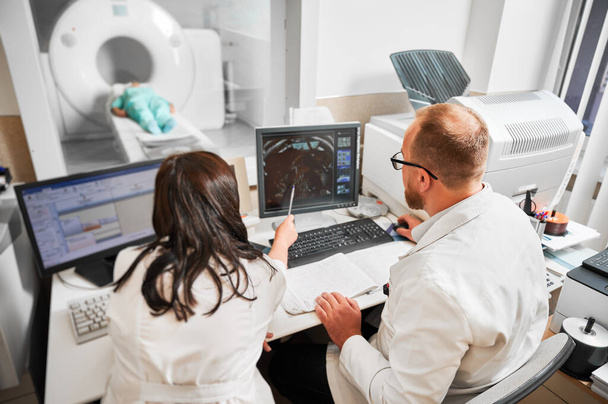 Widok dwóch lekarzy siedzących przy komputerze, badających wyniki rezonansu magnetycznego. Specjaliści ubrani w białe szaty, konsultanci, kobieta wskazana długopisem, pacjent kłamie. Pojęcie medycyny. - Zdjęcie, obraz
