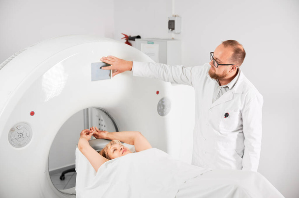 Ιατρική υπολογιστική τομογραφία ή μαγνητική τομογραφία. Αρσενικός ακτινολόγος πατάει το κουμπί MRI για να εξετάσει γυναίκα ασθενή. Έννοια της υγειονομικής περίθαλψης και σύγχρονα διαγνωστικά. - Φωτογραφία, εικόνα