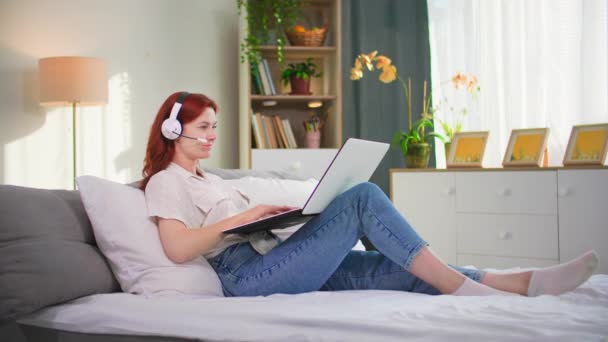 Freiberuflerin, junge Frau arbeitet als Callcenter-Betreiberin zu Hause und kommuniziert mit Headset und Videoanruf am Laptop, während sie zu Hause auf dem Sofa liegt - Filmmaterial, Video