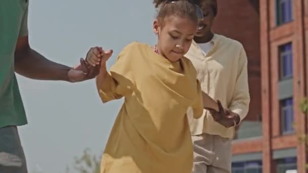 Medium shot of joyous African American mother and father holding hands with little daughter while she riding skateboard for first time outdoors on summer day - Footage, Video