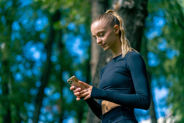Побочный снимок девушки в спортивной одежде, внимательно смотрящей в телефон во время тренировки в зеленом парке - Фото, изображение