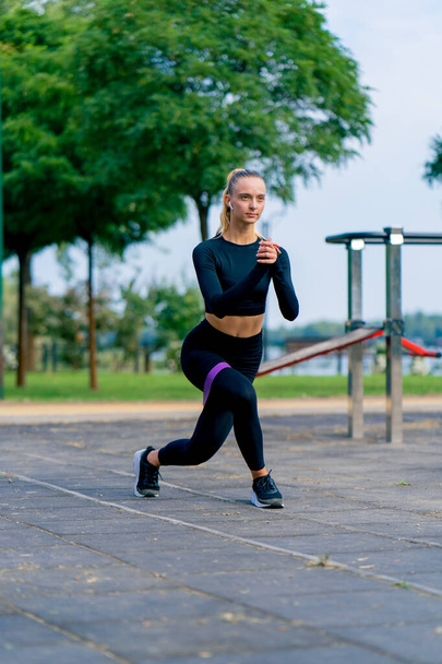 Красивая девушка в спортивной форме выполняет упражнения, чтобы накачать ноги резинкой на спортивной площадке - Фото, изображение