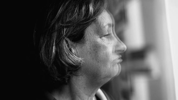 Ängstlich depressive Seniorin mit Großaufnahme, die am Fenster steht und mit ruhiger launischer Selbstbetrachtung nach draußen blickt. Einfarbiges Schwarz-Weiß, beunruhigende Emotionen - Foto, Bild