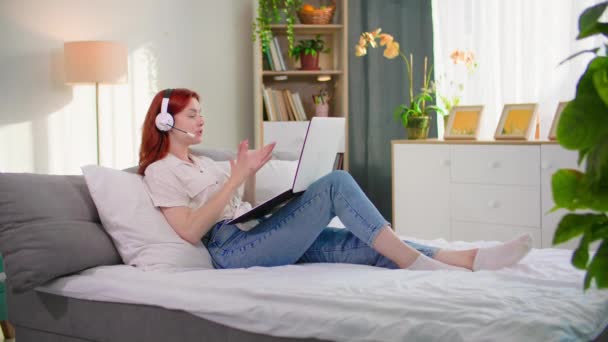 Интернет-общение, молодая женщина разговаривает по видеосвязи с помощью гарнитуры, расслабляясь на диване дома - Кадры, видео