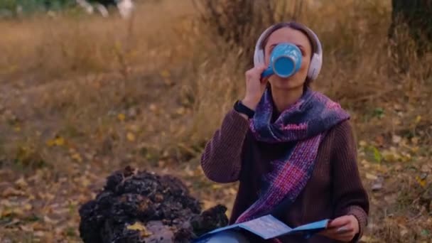 woman with a backpack drinks tea from a cup, dressed in a brown sweater, holds a map in her hands, enjoys sunny weather in the autumn forest, traveler on the precipice, My Real Holiday, MyRealHoliday - Footage, Video