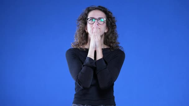 Geïsoleerde jonge vrouw die op een blauwe achtergrond staat en omhoog kijkt met handen tegen elkaar geklemd haar adem inhouden tijdens stressvolle tijden - Video