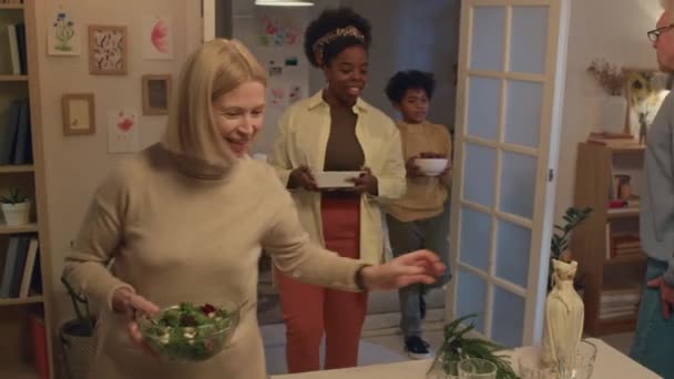 Leden van multi-etnische familie die zelfgemaakte cake, salade en ander eten naar de eettafel dragen in de gezellige woonkamer - Video