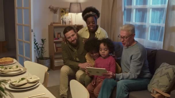 Interkulturelle Mehrgenerationenfamilie schaut sich Videos auf digitalem Tablet an, während sie zusammen auf dem Sofa neben dem festlichen Tisch im Wohnzimmer sitzt - Filmmaterial, Video