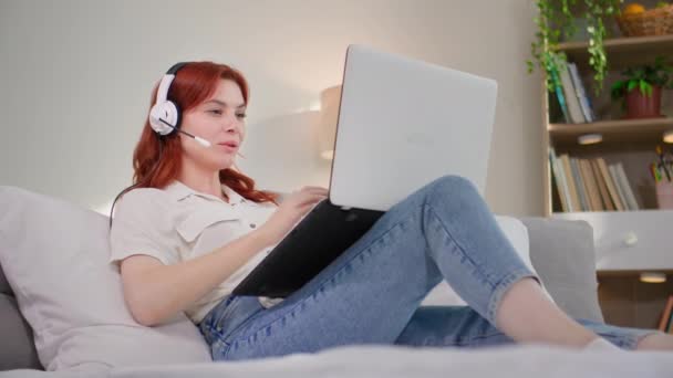 szabadúszó munka, fiatal gyönyörű nő dolgozik online call center és kommunikál az ügyféllel segítségével headset és laptop, miközben fekszik a kanapén a szobában - Felvétel, videó