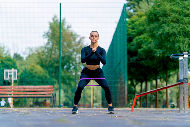 Bella ragazza in uniforme sportiva esegue esercizi per pompare le gambe con un elastico su un parco giochi sportivo di strada - Foto, immagini