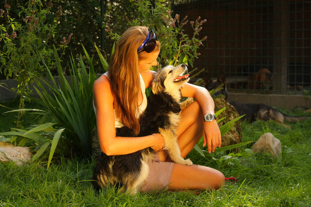 ブロンドの髪を持つ美しい女が保持している愛情を込めて野良犬と緑の草の裏庭の庭で座っている彼女の腕の中 - 写真・画像
