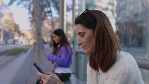 トラムステーションプラットフォームで待機しているスマートフォンを使用する2人の若い女性. 公共交通機関の停留所に座っているセル・スクリーンを見ている大人の通勤女性. シティライフスタイルとテクノロジーコンセプト. - 映像、動画