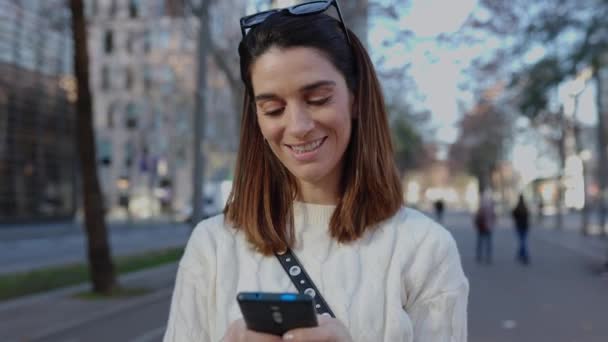 Όμορφη νεαρή ενήλικη γυναίκα χρησιμοποιώντας μια συσκευή smartphone στο δρόμο της πόλης. Millennial γυναίκα σε casual ρούχα γραπτών μηνυμάτων ή βλέποντας περιεχόμενο των μέσων κοινωνικής δικτύωσης πάνω από αστικό υπόβαθρο. Έννοια τρόπου ζωής τεχνολογίας. - Πλάνα, βίντεο