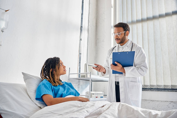 jong Indiaas arts kijken naar Afrikaans-Amerikaanse patiënt liggend in bed in ziekenhuis afdeling, gezondheidszorg - Foto, afbeelding