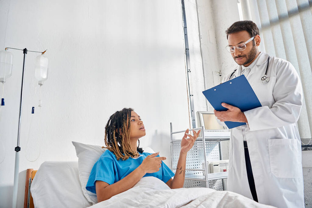 Genç Hintli doktor, Afro-Amerikan hastasına bakıyor. Hastane koğuşunda yatıyor. - Fotoğraf, Görsel