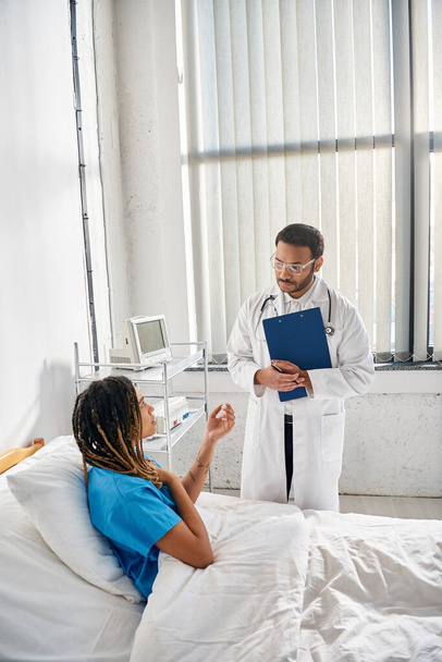 guapo médico indio en gafas hablando con joven afroamericano paciente en el hospital, atención médica - Foto, Imagen