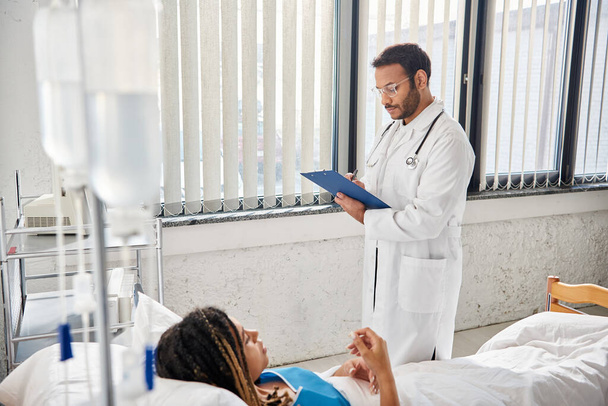 gut aussehender indischer Arzt macht Notizen über afrikanisch-amerikanische Patientin, die auf Krankenhausstation im Bett liegt - Foto, Bild