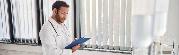 młody indyjski lekarz w okularach i stetoskopie na oddziale szpitalnym robi notatki, opiekę zdrowotną, baner - Zdjęcie, obraz