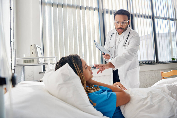 doctora india inteligente hablando con su paciente afroamericana acostada en la cama en la sala de salud - Foto, Imagen
