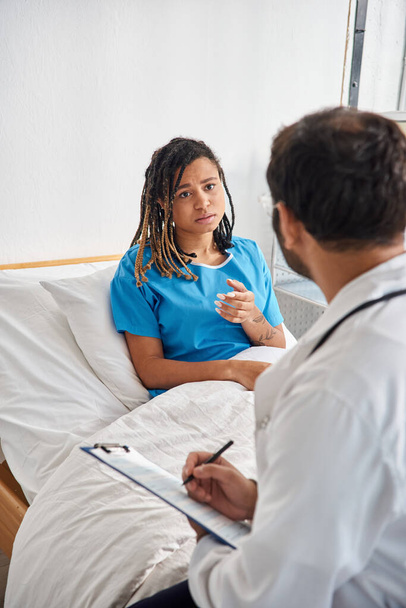atractiva mujer afroamericana enferma acostada en la cama de hospital hablando con su joven médico indio - Foto, Imagen