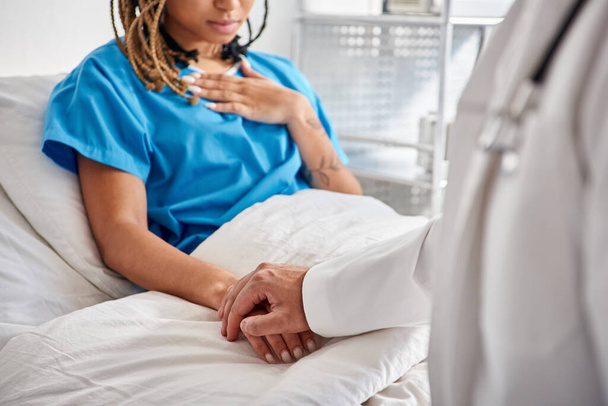vista recortada del joven médico indio sosteniendo su mano de paciente afroamericano acostado en la cama del hospital - Foto, Imagen