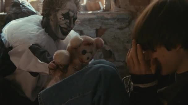 Средний снимок клоунского маньяка, пугающего похищенную девушку с плюшевым мишкой, сидящего на полу - Кадры, видео