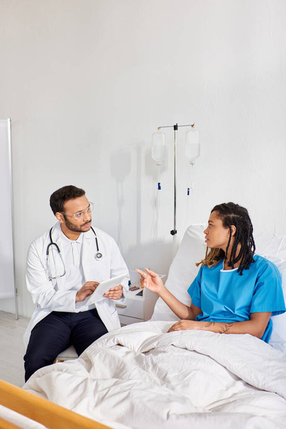 afro-américaine patiente couchée dans un lit d'hôpital parlant à son médecin indien prenant des notes sur tablette - Photo, image